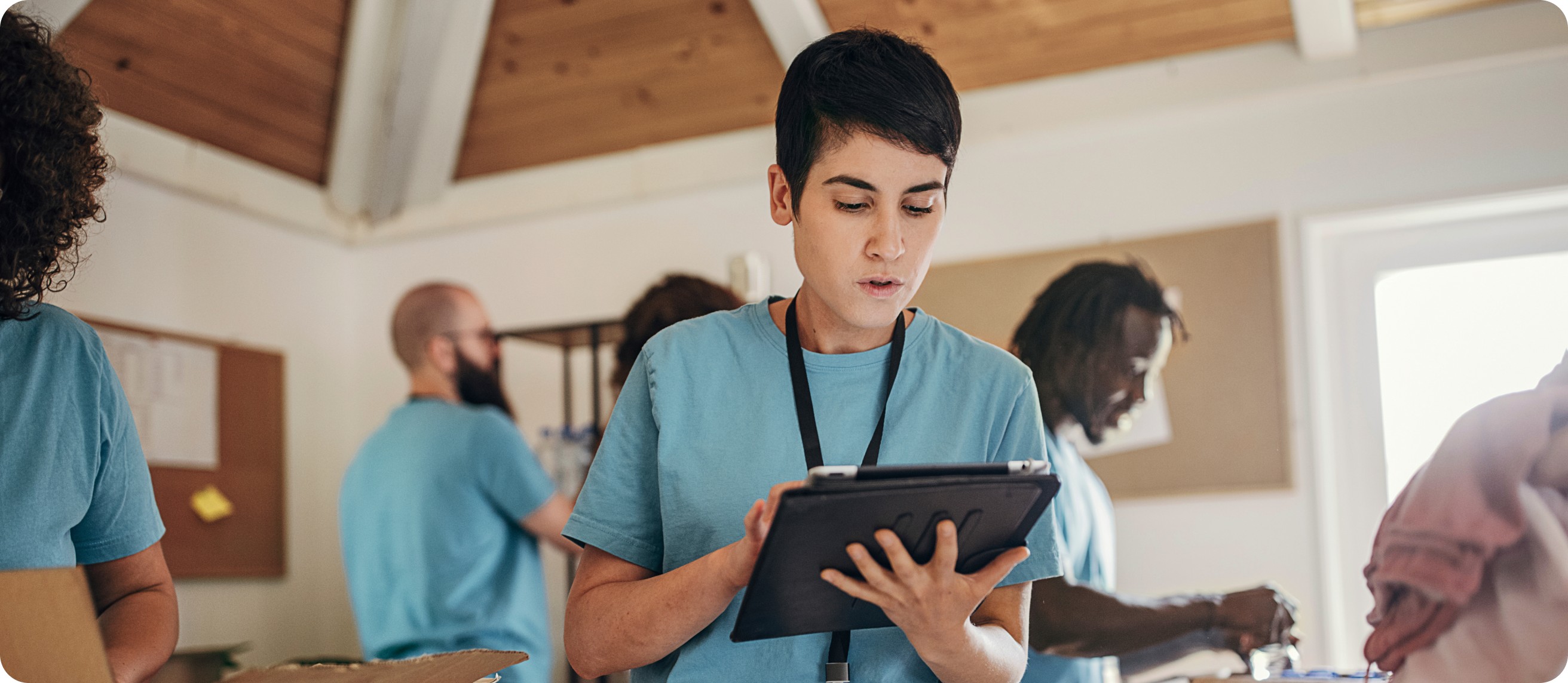 Woman in blue shirt wearing lanyard while looking down at smart device and standing in room with people wearing the same uniform